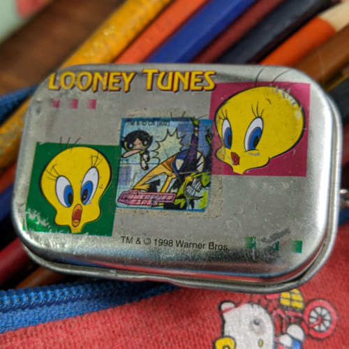 A photograph of a small metal container with the Looney Tunes character Tweety making shocked and happy expressions, as well as a worn out sticker of the Powerpuff Girls. The object is placed on a worn out Hello Kitty bag filled with pencils.